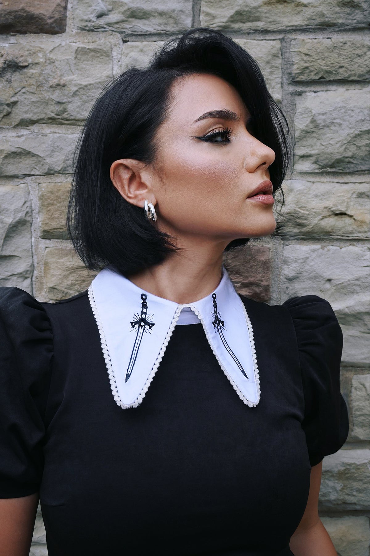 A bold and dramatic black suede Bellatrix Dress with a tight-fitting, short body, oversized puff sleeves, and a prominent white pointed collar with detailed black sword embroidery, perfect for gothic fashion enthusiasts seeking a 1960s-inspired alternative dress that exudes confidence and individuality.