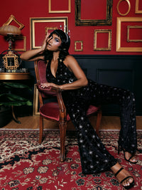Chic and playful Cosmo Jumpsuit in luxurious black velvet with silver starbursts, a halter neckline, and stretchy fabric for comfort, embodying 70s-inspired retro elegance with art deco charm for evening parties and holiday outfits.