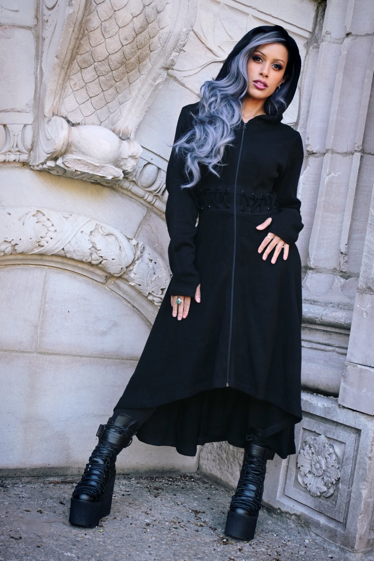 A dramatic full-length black knit cardigan with a structured edge, featuring a fitted bodice, front and back corset-style lacing, zip closure, oversized hood, high-low hem, and thumbholes, made from 100% polyacrylic, perfect for layering or wearing solo for a moody, gothic-inspired look.