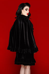 Elegant black stretch velvet cape with a one-size drape that flatters all body types, showcasing a soft, comfortable fit and sophisticated design. Accented with a velvet ribbon tie and ruffled trim, the Hecate Cape brings a romantic and dramatic touch to any wardrobe. Ideal for layering over dresses, tops, or casual outfits, this luxurious cape combines comfort, style, and timeless gothic-inspired aesthetics.