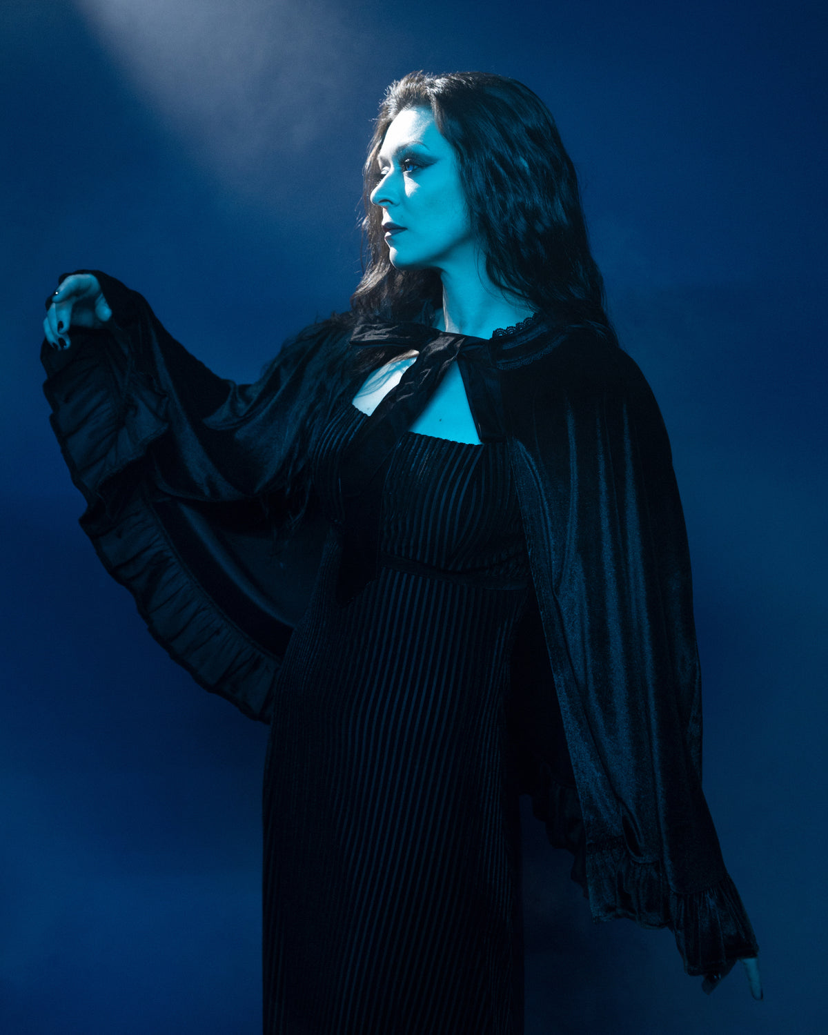 The Hecate Cape is a black velvet statement piece, crafted from soft stretch fabric to provide a comfortable, flowing silhouette for one-size versatility. Featuring a delicate velvet ribbon tie and ruffle detailing, this cape adds a touch of drama and elegance to layered outfits. Perfect for gothic fashion, evening wear, or sophisticated everyday looks.