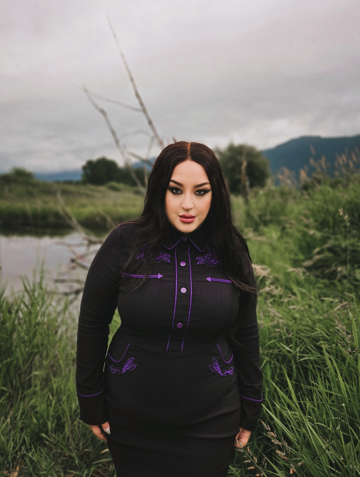 Sophisticated black and purple wiggle dress in stretchy bengaline, featuring long piped sleeves with decorative faux snap cuffs, front snaps for a seamless appearance, and elaborate rose and thorn embroidery across the collar, bust, hips, and cuffs, accented with white piping to complete the refined spring look.