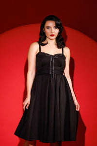 Jessica Louise Love Bites Swing Dress in a luxurious heavyweight black poly blend, featuring a classic fit-and-flare silhouette with a sweetheart bustline, playful ruffled details, and a charming ribbon bow accent, designed with enlarged cup sizing for an effortless, confident fit, side seam pockets for practicality, ruched backing for easy wear, and adjustable straps for a personalized, flattering look.