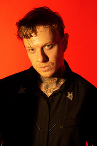 A structured black bengaline western shirt designed with a gothic edge, the Wayne Shirt blends modern alternative fashion with classic cowboy style. Crafted from durable yet form-fitting black bengaline fabric, it features sleek vegan leather piping along the yoke and seams, matte black snap buttons for easy closure, and removable silver collar tips that add a touch of metallic flair. Perfect for those seeking a dark western aesthetic.