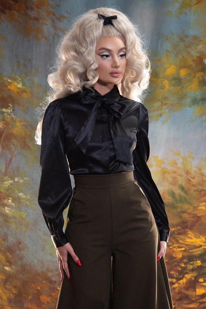 A blond pinup model posing against a yellow and blue painted background while wearing the Angela Blouse in black and brown trousers.