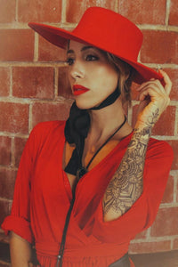 Pinup model posing against a red brick wall wearing a red, wide brimmed, wool hat.