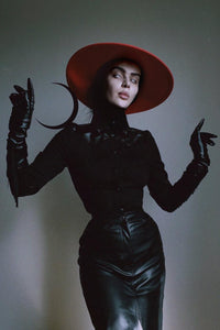 Goth model posing against a white wall wearing a red, wide brimmed, wool hat.