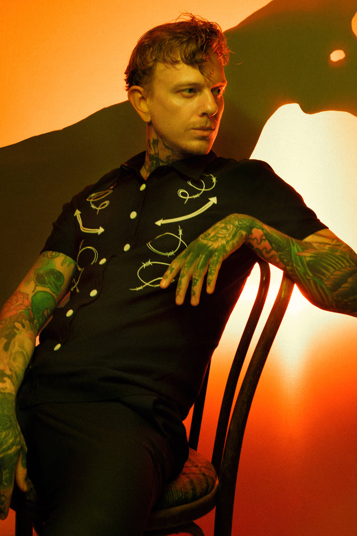 A black fitted short-sleeve men’s shirt crafted from premium high-stretch Bengaline fabric, designed for both comfort and lasting shape retention. The chest is adorned with custom embroidered barbed wire and arrow motifs that blend western grit with a gothic edge, creating a striking statement look. Matte snap buttons line the front for a rugged, durable finish that complements the bold aesthetic and enhances the shirt’s structured silhouette.