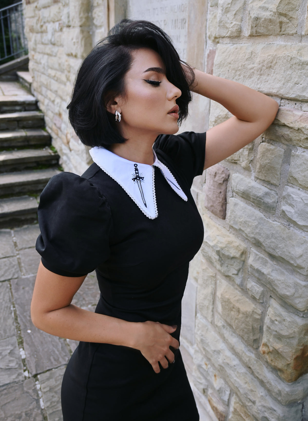 A standout gothic-inspired Bellatrix Dress with a fitted black suede body, exaggerated puff sleeves, and a large white pointed collar embellished with black sword embroidery, perfect for alternative fashion lovers who appreciate 1960s-inspired retro designs and bold, structured statement dresses.