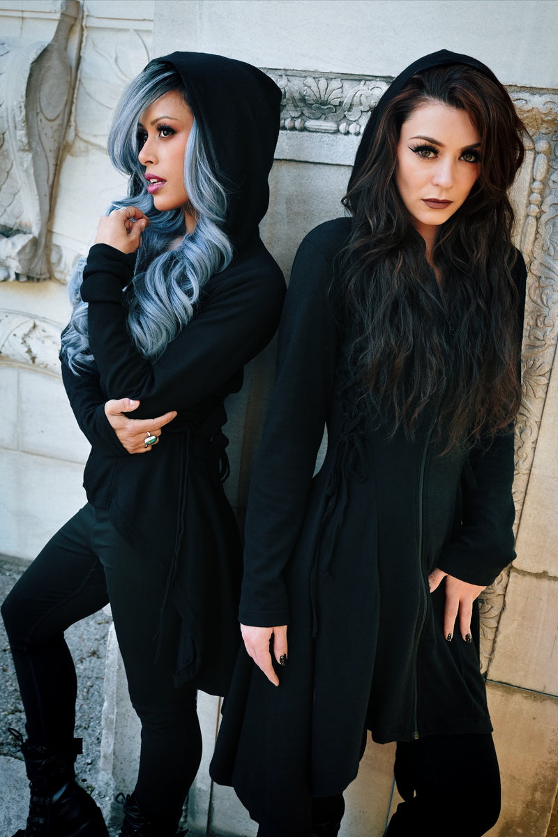 Black zip-up hoodie from Heartless with a soft 100% polyester construction, corset-style side lacing, adjustable back ties, high-low hem, and thumbholes, offering a fitted silhouette, oversized hood, and versatile layering option for casual or statement outfits.