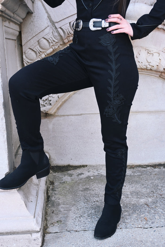 Black stretch denim Dallas Pants, high-waisted and form-fitting, designed with tonal black rose and thorn embroidery along both legs. Includes functional belt loops and a back zipper closure, combining edgy, dark romantic style with comfortable, structured wear.