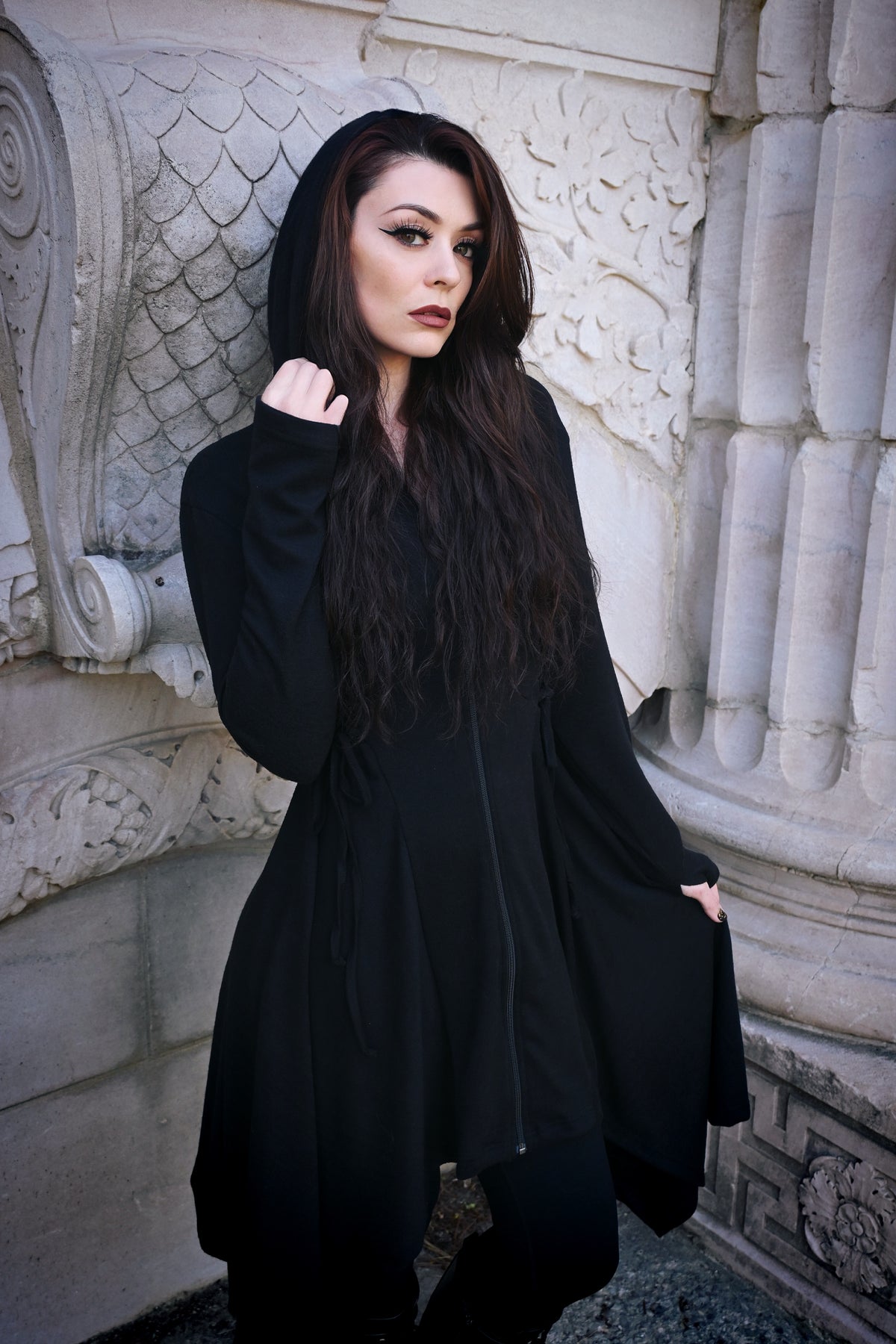 The Emerson Cardigan in black polyester from Innocent Lifestyle, offering a dramatic, mid-length fit with hood, zip-up closure, corset lacing details at sides and back, and a flowing handkerchief hem perfect for layering over casual or evening outfits.