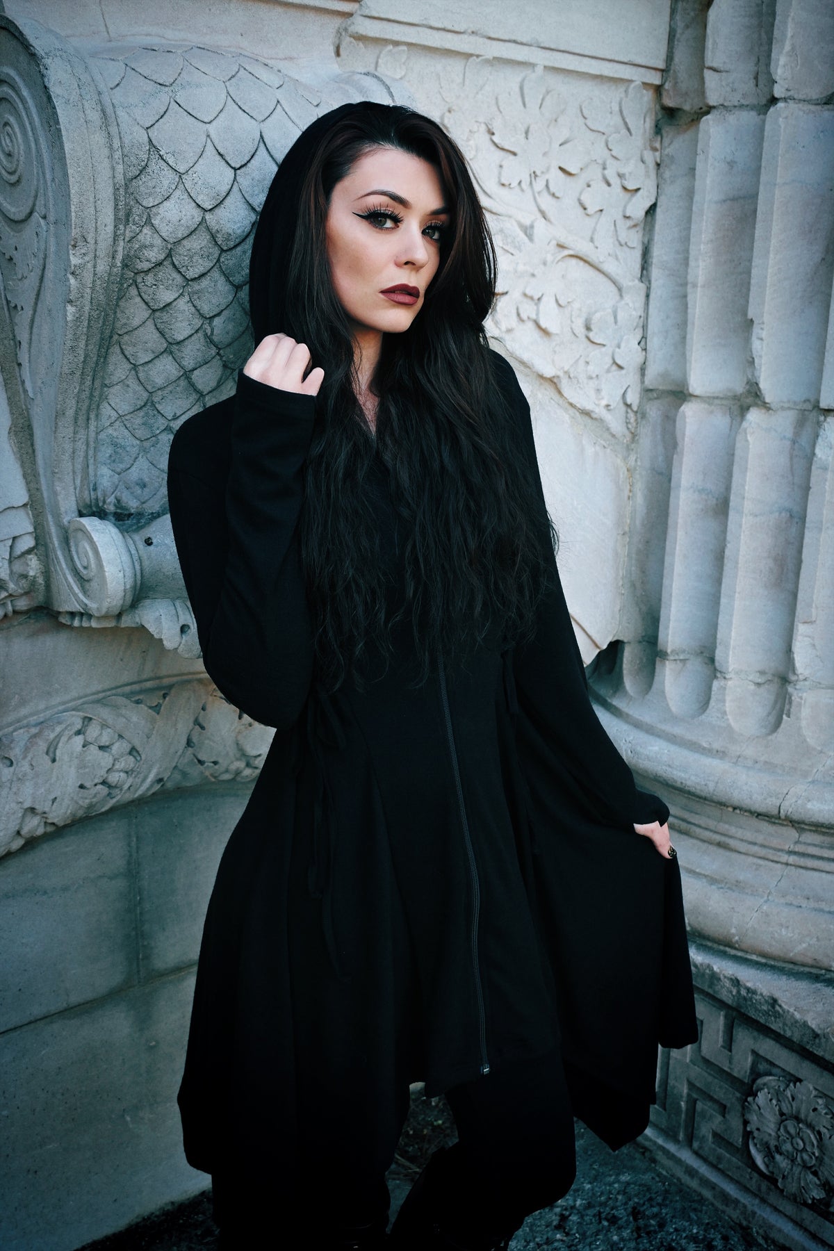 Lightweight black Emerson Cardigan by Innocent Lifestyle, designed with soft drape, mid-length fit, zip-up front, elongated hood, and decorative corset lacing at the sides and back, finished with an asymmetrical handkerchief hem for gothic-inspired layering.