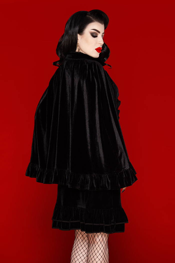 Elegant black stretch velvet cape with a one-size drape that flatters all body types, showcasing a soft, comfortable fit and sophisticated design. Accented with a velvet ribbon tie and ruffled trim, the Hecate Cape brings a romantic and dramatic touch to any wardrobe. Ideal for layering over dresses, tops, or casual outfits, this luxurious cape combines comfort, style, and timeless gothic-inspired aesthetics.