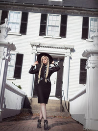 Woman in a black outfit and hat standing in front of a white house with black shutters.