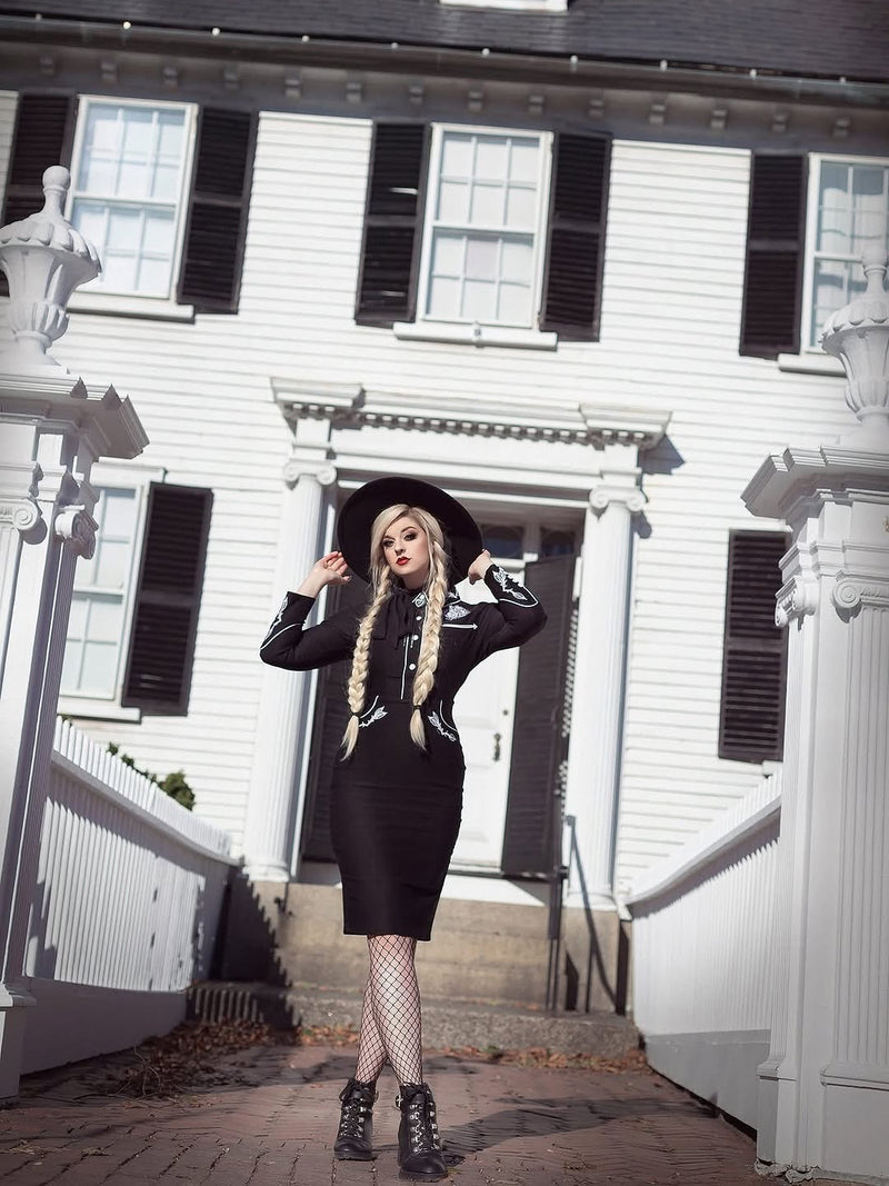 Woman in a black outfit and hat standing in front of a white house with black shutters.