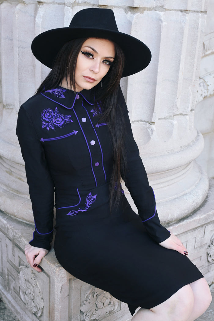 A black and purple June Dress crafted from stretchy bengaline fabric, featuring long piped sleeves with faux snap cuffs, fixed front snaps for a seamless design, and intricate rose and thorn embroidery on the collar, cuffs, bust, and hips, accented by white piping for a sophisticated spring wiggle dress look.