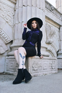 Elegant June Dress in black and purple with form-fitting stretchy bengaline material, showcasing long piped sleeves, decorative faux snap cuffs, front snaps designed for a smooth silhouette, and delicate rose and thorn embroidery throughout the collar, bust, hips, and cuffs, finished with contrasting white piping.