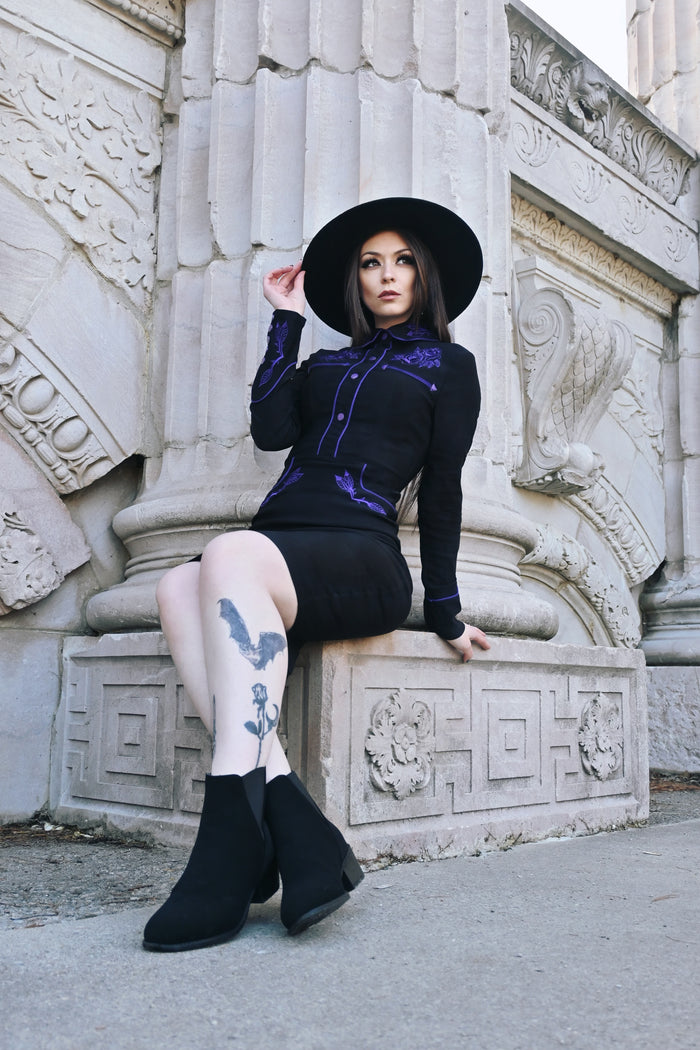 Elegant June Dress in black and purple with form-fitting stretchy bengaline material, showcasing long piped sleeves, decorative faux snap cuffs, front snaps designed for a smooth silhouette, and delicate rose and thorn embroidery throughout the collar, bust, hips, and cuffs, finished with contrasting white piping.
