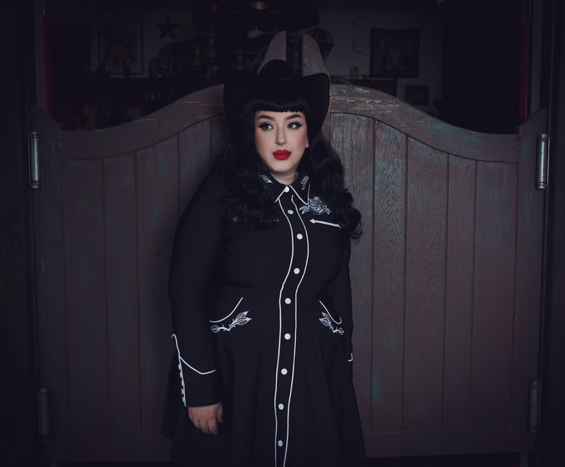 Black and white June Dress featuring a feminine skater skirt design, intricate rose and thorn embroidery along the collar, bust, hips, and cuffs, elegant white piping accents, long piped sleeves with faux snap cuffs, and deep, practical pockets, combining whimsical charm, chic detailing, and everyday versatility.