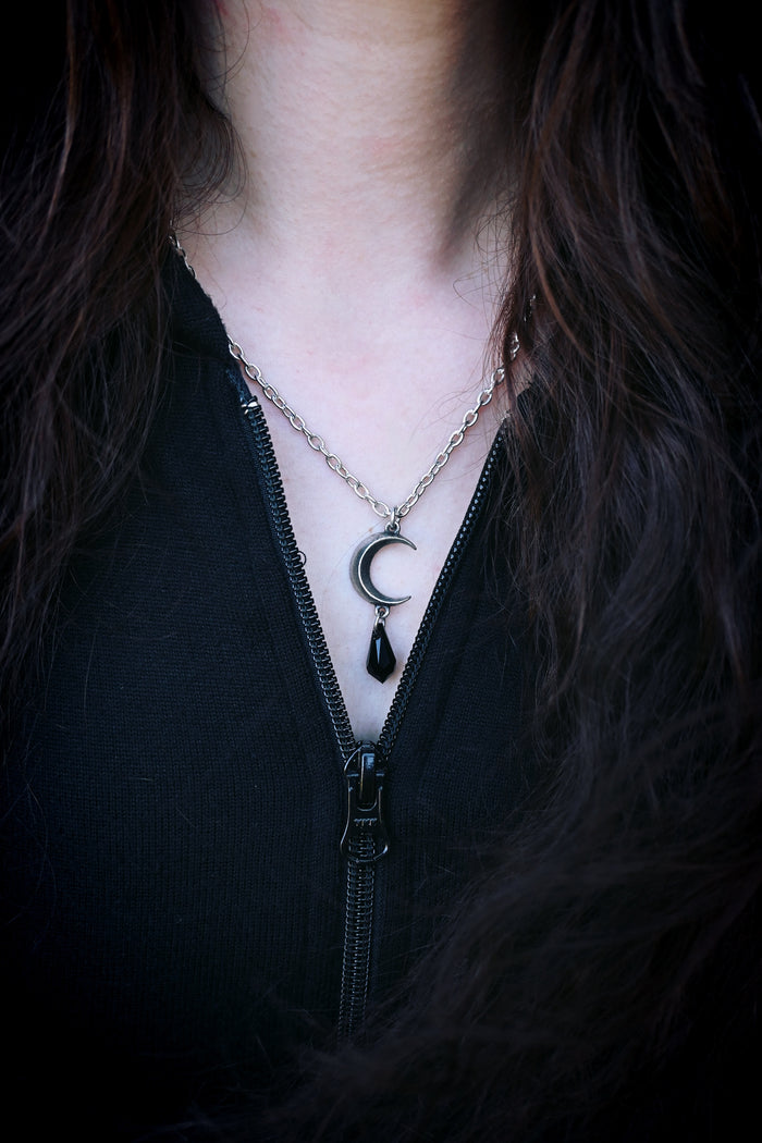 This pendant offers a dramatic blend of celestial symbolism and refined craftsmanship—crafted in English pewter, the moon-shaped top supports a faceted jet-black crystal drop that catches light and attention. Designed with adjustable chain length and night-inspired narrative (“the Goddesses' tears cleave the night asunder”), the piece serves as both jewellery and storytelling accessory.