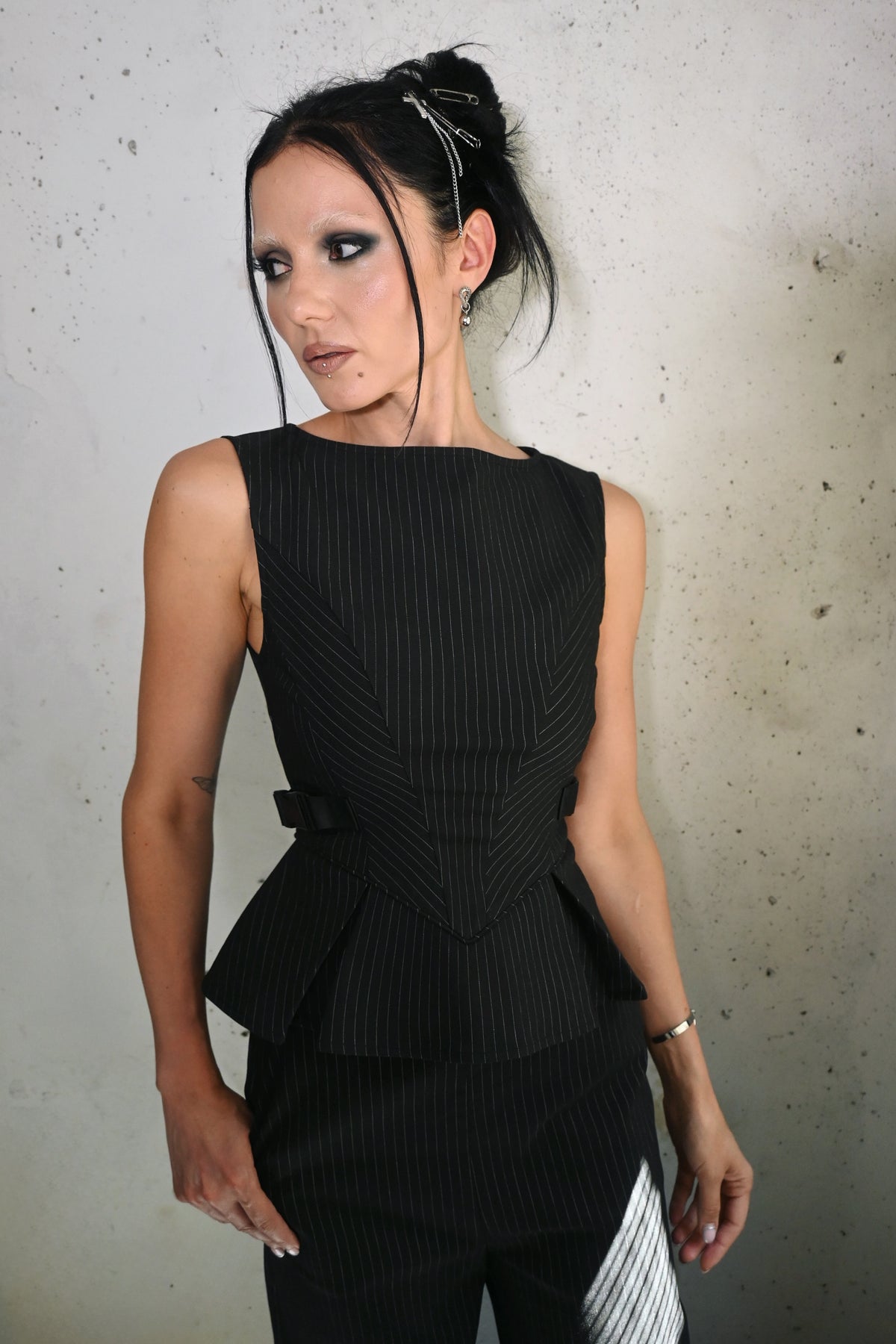 A woman modeling a black sleeveless pinstriped peplum top and matching pants against a textured concrete wall.