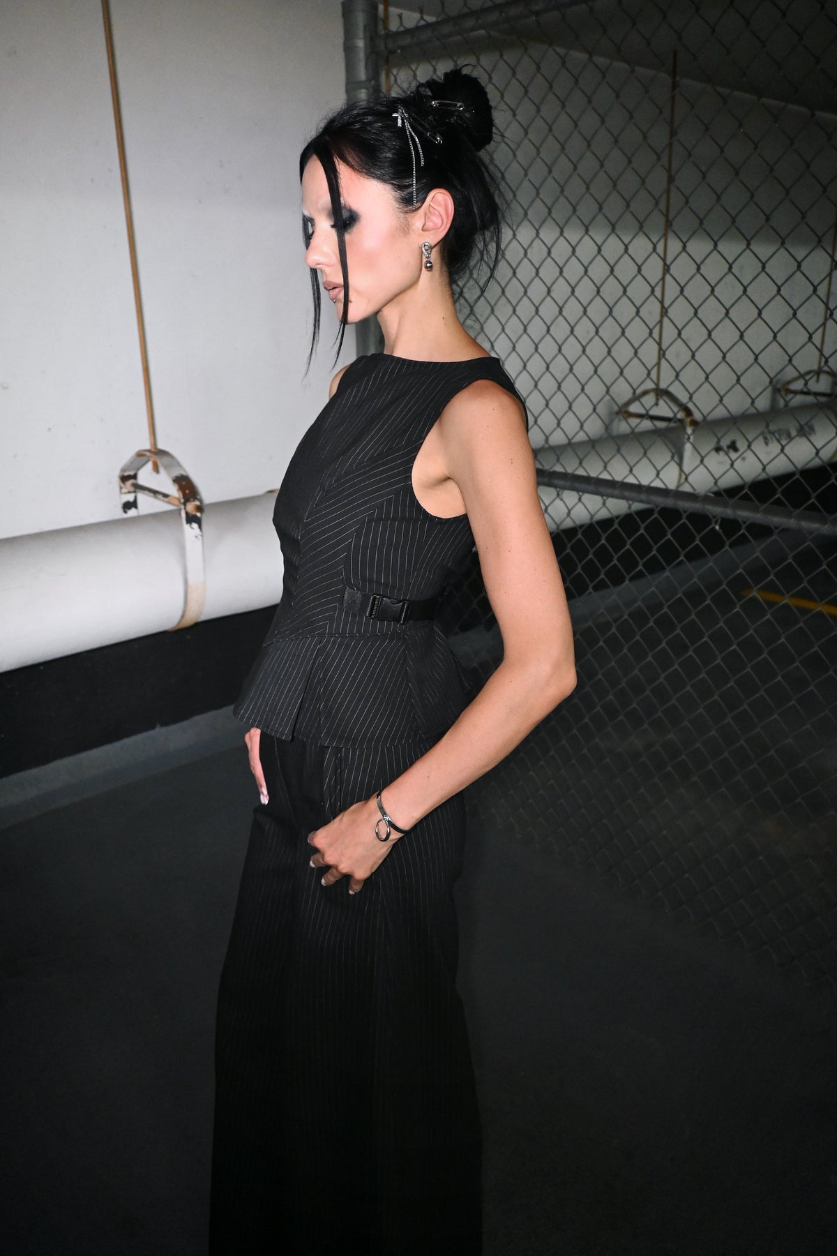 A woman wearing a sleeveless pinstripe top with a peplum waist, standing in a dimly lit parking structure with a chain-link fence in the background.