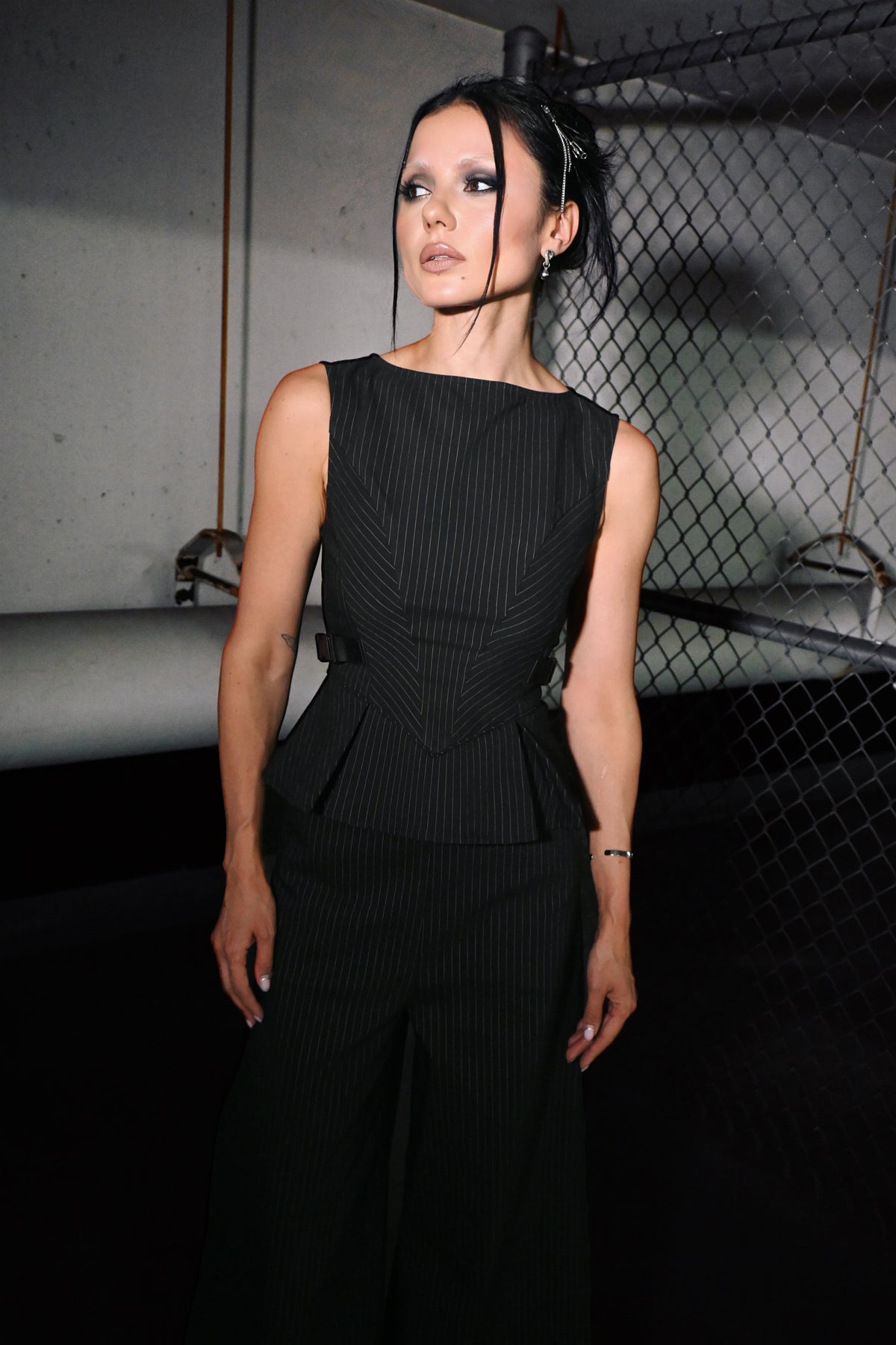 A woman modeling a black sleeveless pinstriped top with a structured peplum waist, set against a dark industrial background with a chain-link fence.