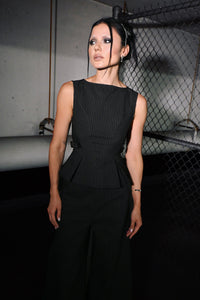 A woman modeling a black sleeveless pinstriped top with a structured peplum waist, set against a dark industrial background with a chain-link fence.