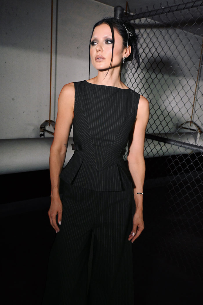 A woman modeling a black sleeveless pinstriped top with a structured peplum waist, set against a dark industrial background with a chain-link fence.