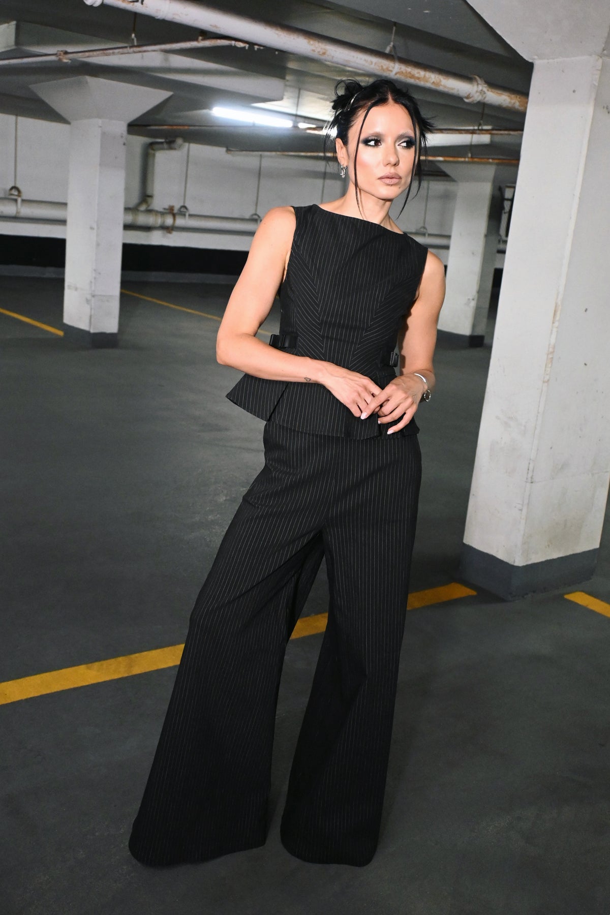 A woman wearing a black sleeveless pinstripe top with a peplum waist and wide-leg palazzo pants, standing in a concrete parking garage.
