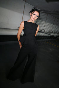 A model wearing a black pinstripe sleeveless peplum top and matching wide-leg trousers, standing in a dimly lit concrete structure with a chain-link fence in the background.