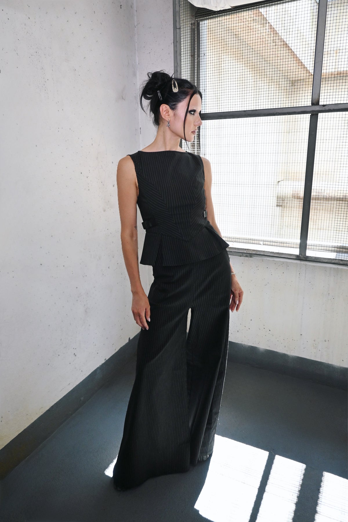 A model stands in a minimalist room with concrete walls and a large window, wearing a black sleeveless peplum top and matching wide-leg trousers.