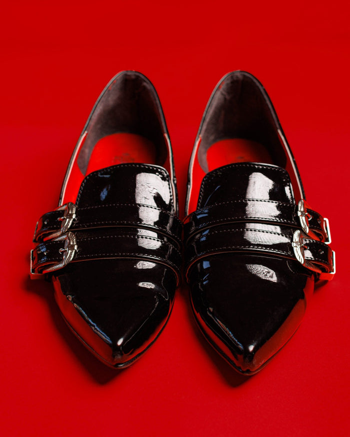 Front view of the Antonella flat shoes in patent black against a red background.