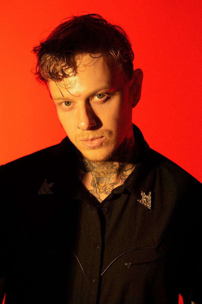 A structured black bengaline western shirt designed with a gothic edge, the Wayne Shirt blends modern alternative fashion with classic cowboy style. Crafted from durable yet form-fitting black bengaline fabric, it features sleek vegan leather piping along the yoke and seams, matte black snap buttons for easy closure, and removable silver collar tips that add a touch of metallic flair. Perfect for those seeking a dark western aesthetic.