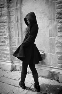 A black hooded gothic dress designed to provide warmth, comfort, and an elegant silhouette. Crafted from 100% heavyweight 400 GSM cotton with a soft fleece lining, the dress features a fitted waist and full skater-style skirt for a flattering shape. A deep hood and oversized pockets add cozy functionality, while the gothic cross zipper pull provides a subtle statement detail.