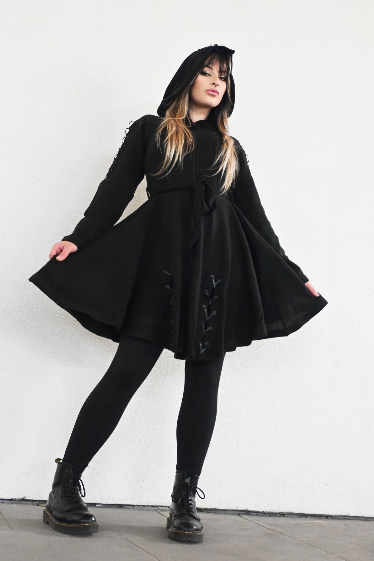 The Fialka Cardigan is a longline black knit cardigan that combines dark romantic elegance with whimsical elfish styling. It features an oversized pointed hood with a continuous row of lace-up grommet detailing running down the center of the back. Corset-style eyelet lacing decorates the outer sleeves and continues along the front hemline, adding an intricate alternative touch. A front zipper and matching tie belt allow the flared silhouette to be adjusted and cinched for a more defined waist.