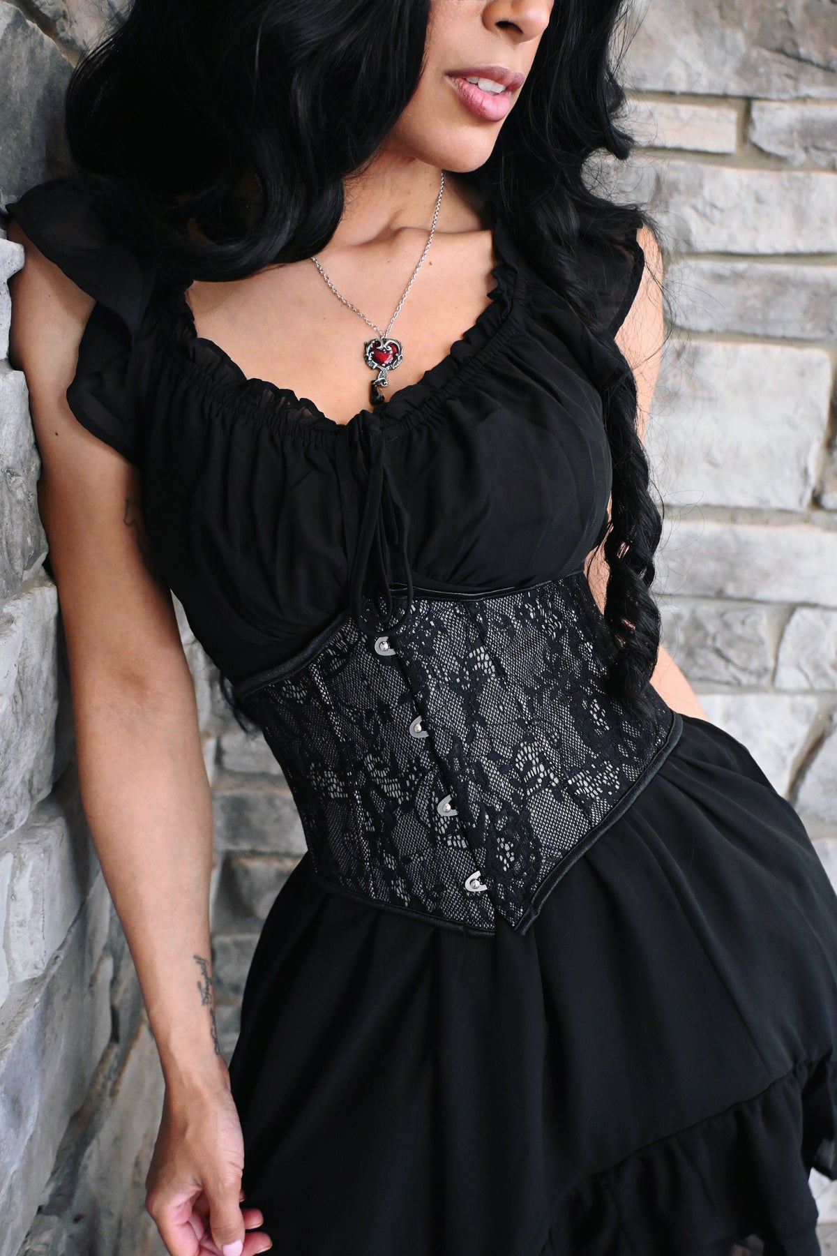 An elegant black lace waspie belt designed to create a dramatic hourglass silhouette. The structured shape cinches the waist, while delicate floral lace layered over a matching lining adds a romantic gothic touch. Decorative hook closures run along the front, and the pointed hem creates a flattering Victorian-inspired finish. Perfect for wearing over dresses, blouses, or flowing tops.