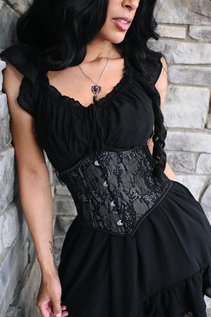 An elegant black lace waspie belt designed to create a dramatic hourglass silhouette. The structured shape cinches the waist, while delicate floral lace layered over a matching lining adds a romantic gothic touch. Decorative hook closures run along the front, and the pointed hem creates a flattering Victorian-inspired finish. Perfect for wearing over dresses, blouses, or flowing tops.