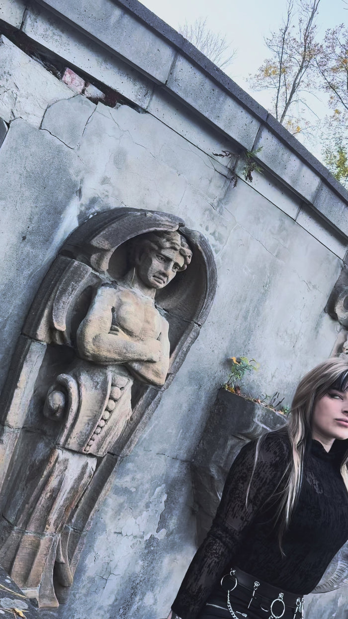 Cropped, straight-leg black pants crafted from a durable cotton and elastane blend. Designed with a mid-rise waist, these gothic-inspired trousers feature silver zip-closure back pockets, matte strap accents on the thighs, and long zippers running up the outer legs. The design is punctuated with metal D-rings and zipper accents.