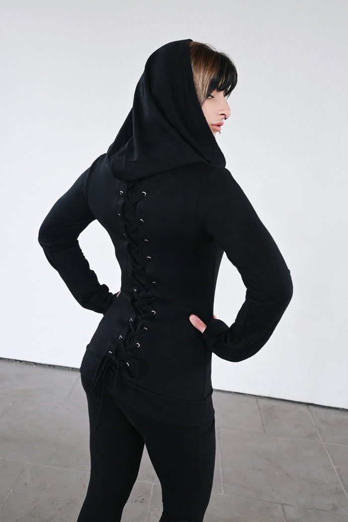 A dramatic longline tunic top featuring a convertible neckline and adjustable corset-inspired back detailing. Crafted from 100% polyester knit, the Rest Top can be styled with the neckline drawn upward as an oversized hood or worn open as a loose, cascading cowl neck. The entire length of the back is decorated with metal eyelets and corset lacing, allowing the wearer to adjust the fit and define the waist. Extra-long sleeves complete the relaxed, oversized silhouette.