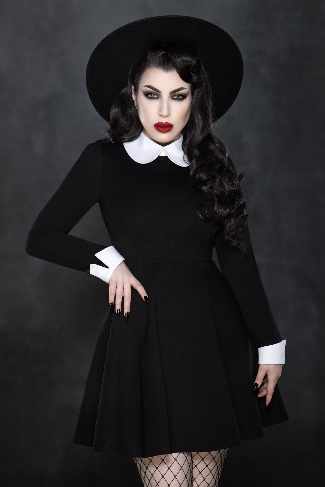 Model posing against grey background wearing Addams Dress, black hat and fishnets. 