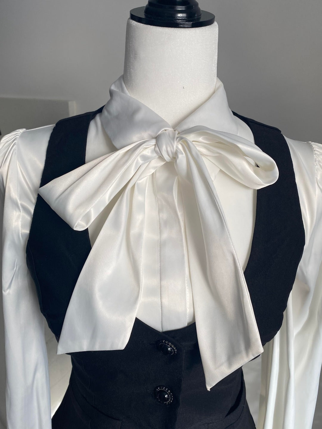 Ribbon details of the Angela Blouse in white with a black vest on top on a white bust mannequin against a white wall.