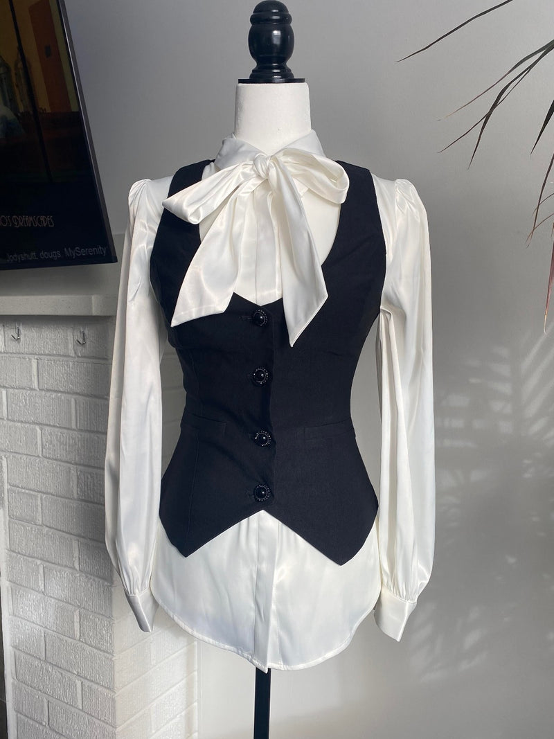 The Angela Blouse in white with a black vest on top on a white bust mannequin again a white wall.