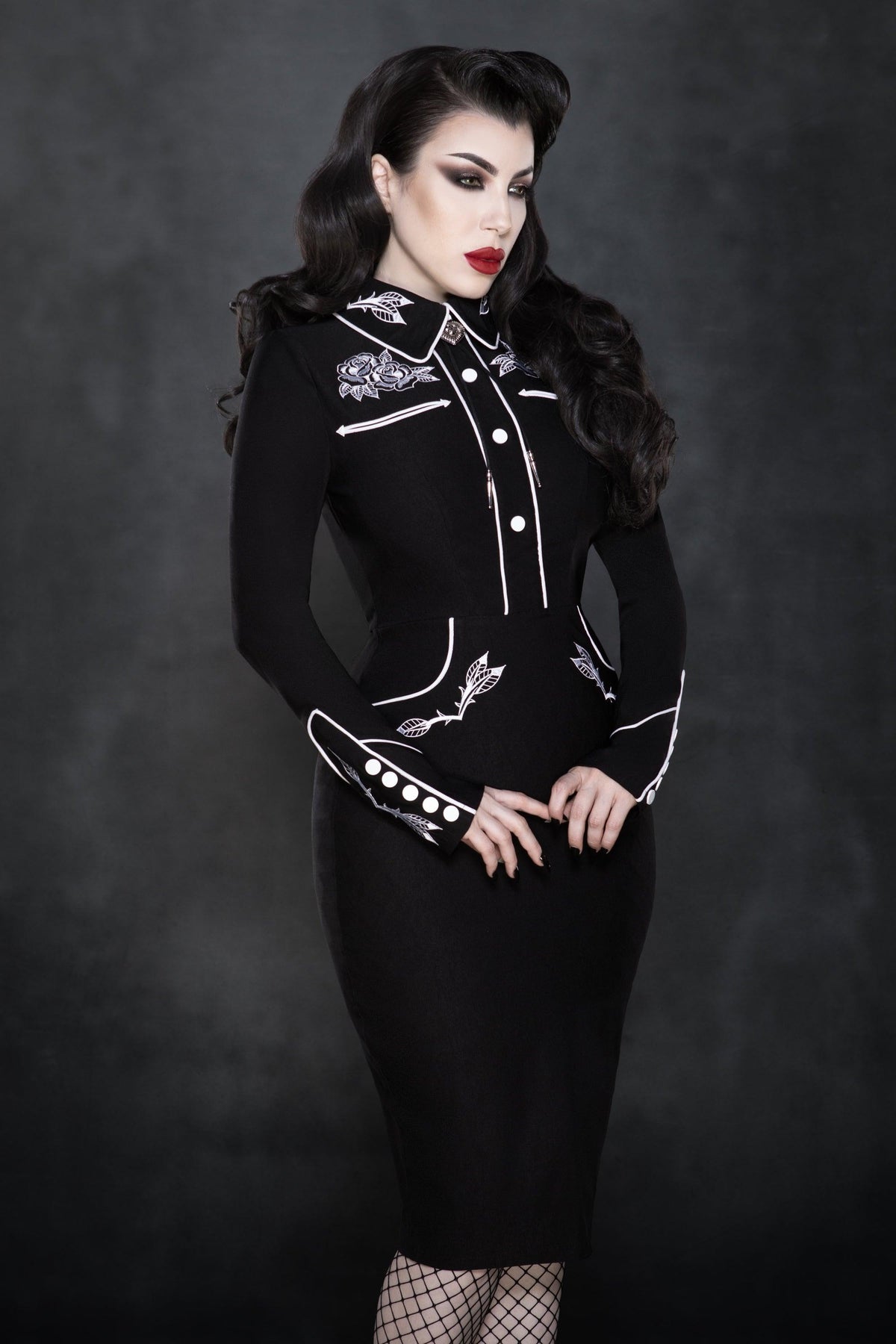 A vintage-inspired black and white stretch bengaline wiggle dress featuring elegant white piping and intricate rose and thorn embroidery on the collar, cuffs, bust, and hips. Designed with long piped sleeves, faux snap cuffs, and a smooth fitted silhouette, this timeless June Carter–inspired piece blends classic charm with modern sophistication — perfect for a statement spring look.