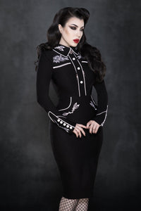 A vintage-inspired black and white stretch bengaline wiggle dress featuring elegant white piping and intricate rose and thorn embroidery on the collar, cuffs, bust, and hips. Designed with long piped sleeves, faux snap cuffs, and a smooth fitted silhouette, this timeless June Carter–inspired piece blends classic charm with modern sophistication — perfect for a statement spring look.