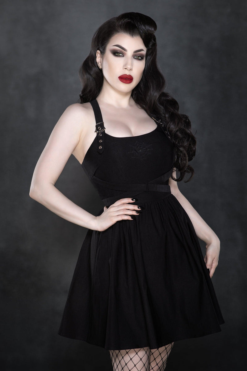 A sleek black skater dress crafted in a comfortable Bengaline fabric, featuring a cross-front strap design that draws the eye upward, a gently flared A-line silhouette that adds glamour and movement, subtle front embroidery details, and distinctive webbed buckle accents at the waist for an edgy yet refined finish.