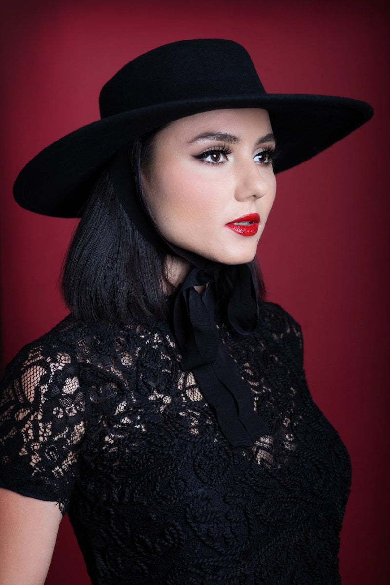 A pinup model posing against a red backdrop while wearing a black, wide brim American Gothic hat.