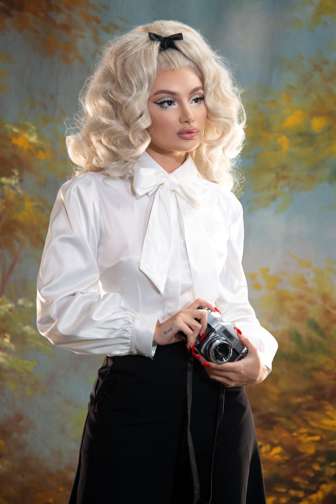 A blond pinup model posing against a yellow and blue painted background while wearing the Angela Blouse in white and black trousers with an old camera on her hand.