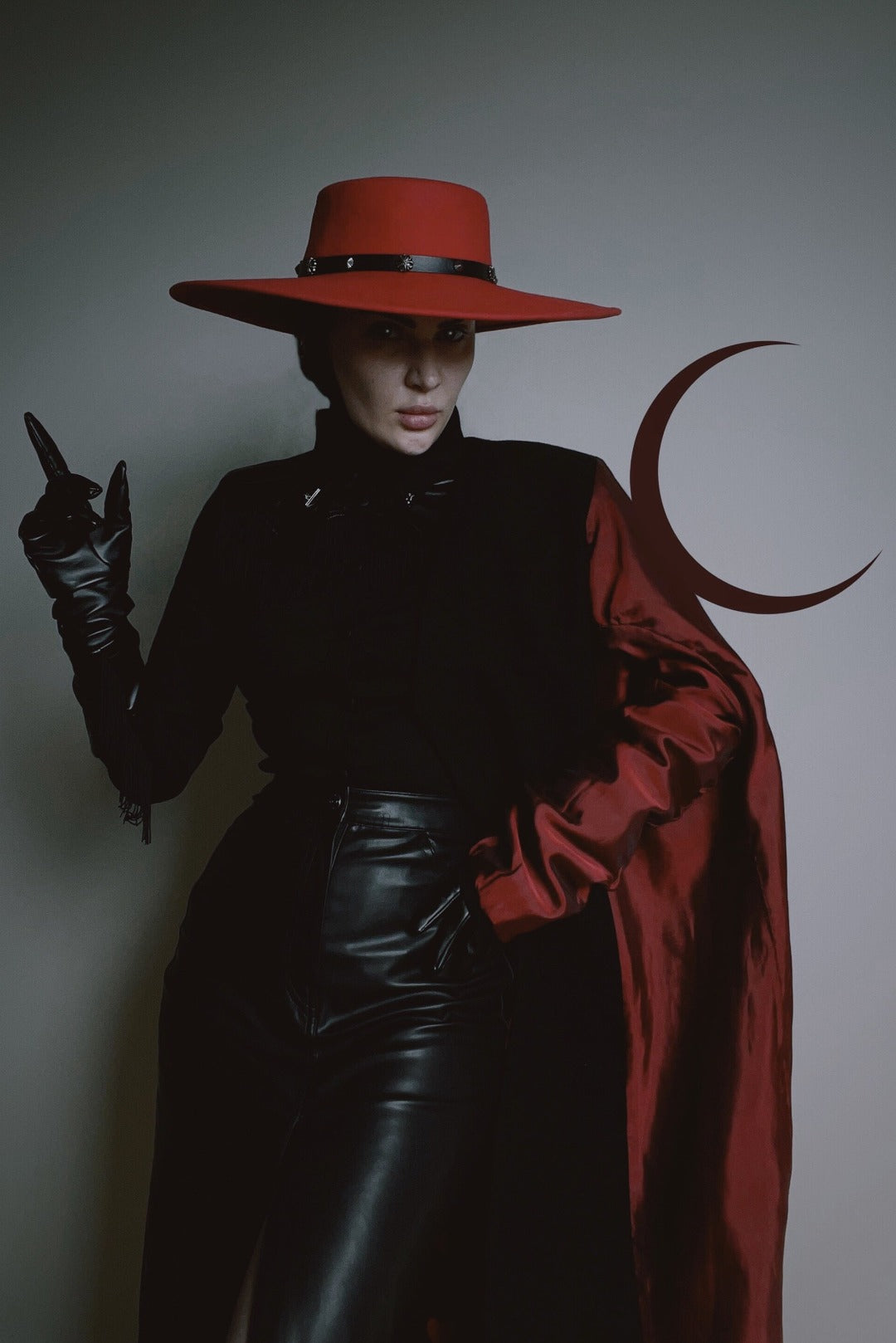 Goth model posing against a white wall wearing a red, wide brimmed, wool hat.